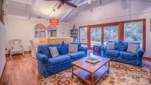 A spacious living room with a high ceiling, a ceiling fan, and large windows. There are two blue sofas with striped pillows, a wooden coffee table, and a patterned rug.