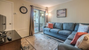 A living room with a sofa, a clock, a television, and a door leading to a balcony.