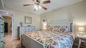 A bedroom with a floral bedspread, a ceiling fan, a dresser, a nightstand, and a lamp.