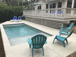 Une piscine avec une terrasse, deux chaises bleues et un espace abrité avec des parasols.
