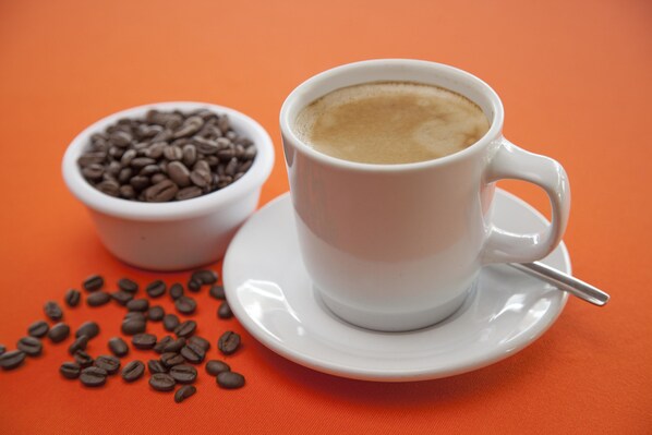 A white cup of coffee with a spoon on a saucer, a bowl of coffee beans, and scattered beans on an orange surface.