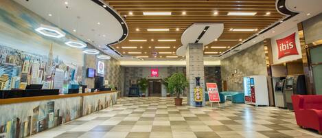 A hotel lobby with a checkered floor, a reception desk, a large potted plant, a vending machine, and a red seating area.