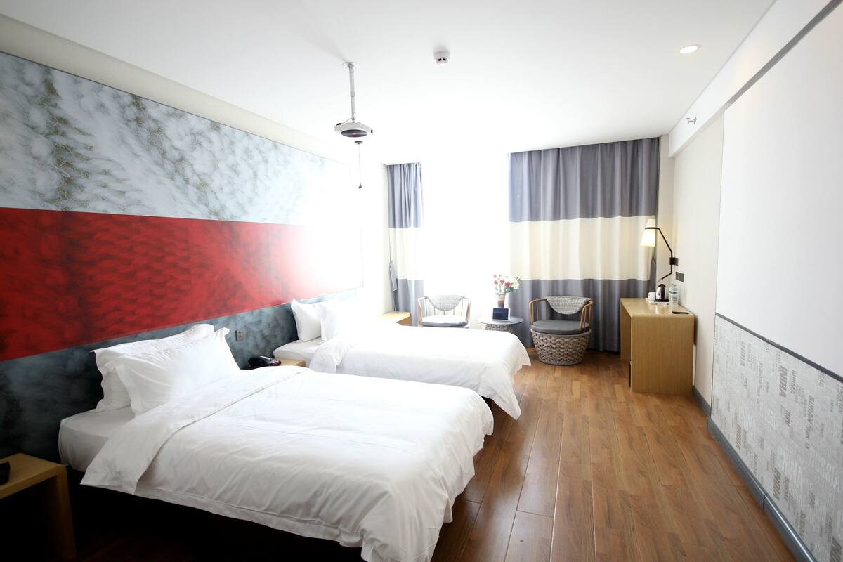 A hotel room with two beds, a small table, a chair, a nightstand, and a wall with a red and grey design.