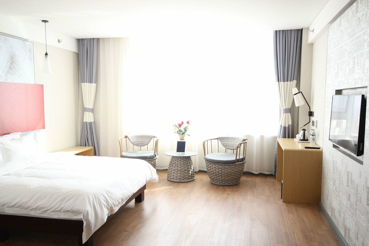 A hotel room with a bed, two wicker chairs, a small table with a vase of flowers, a bedside table with a lamp, and a TV on the wall.