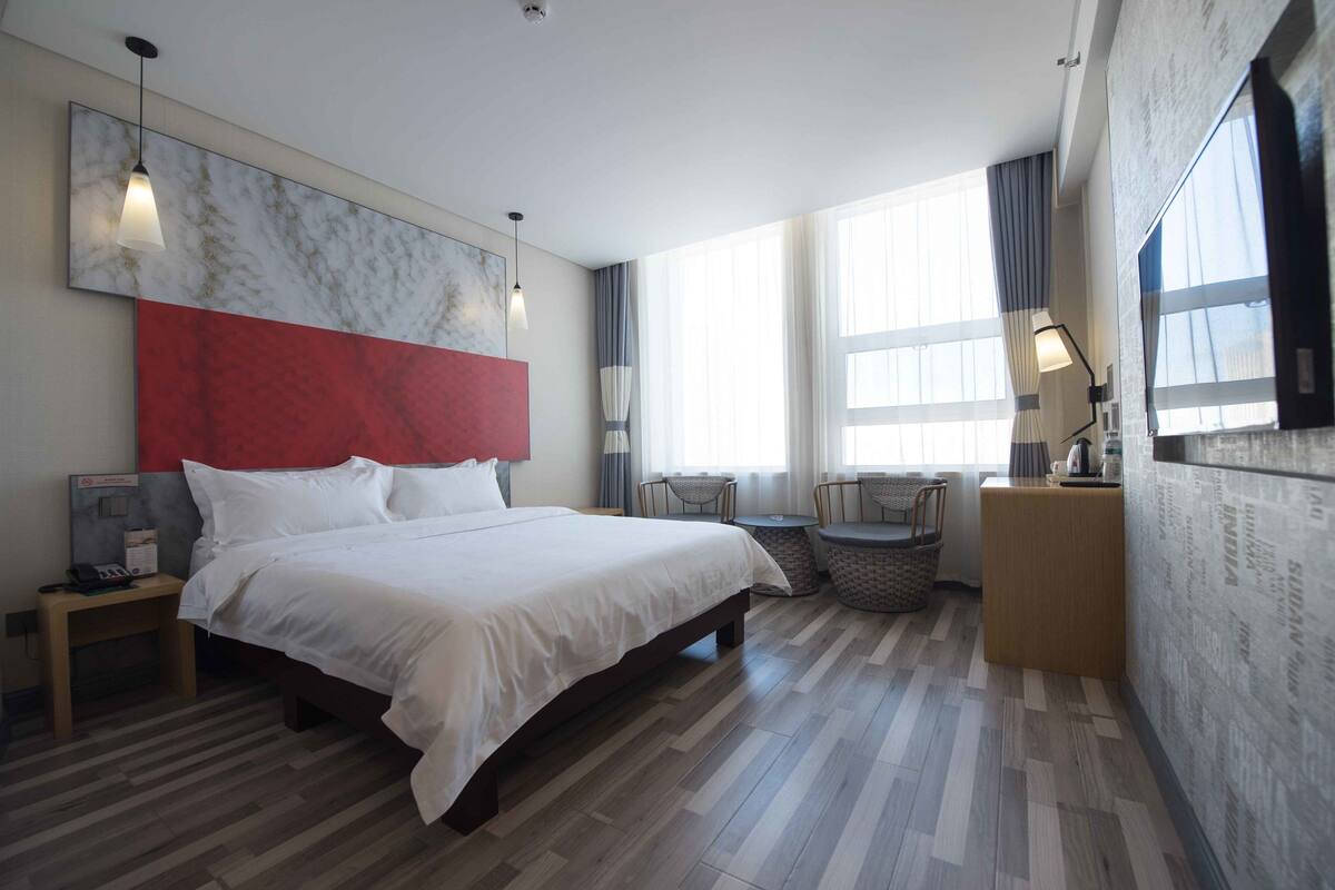 A hotel room with a large bed, a small table, a chair, and a nightstand with a lamp.