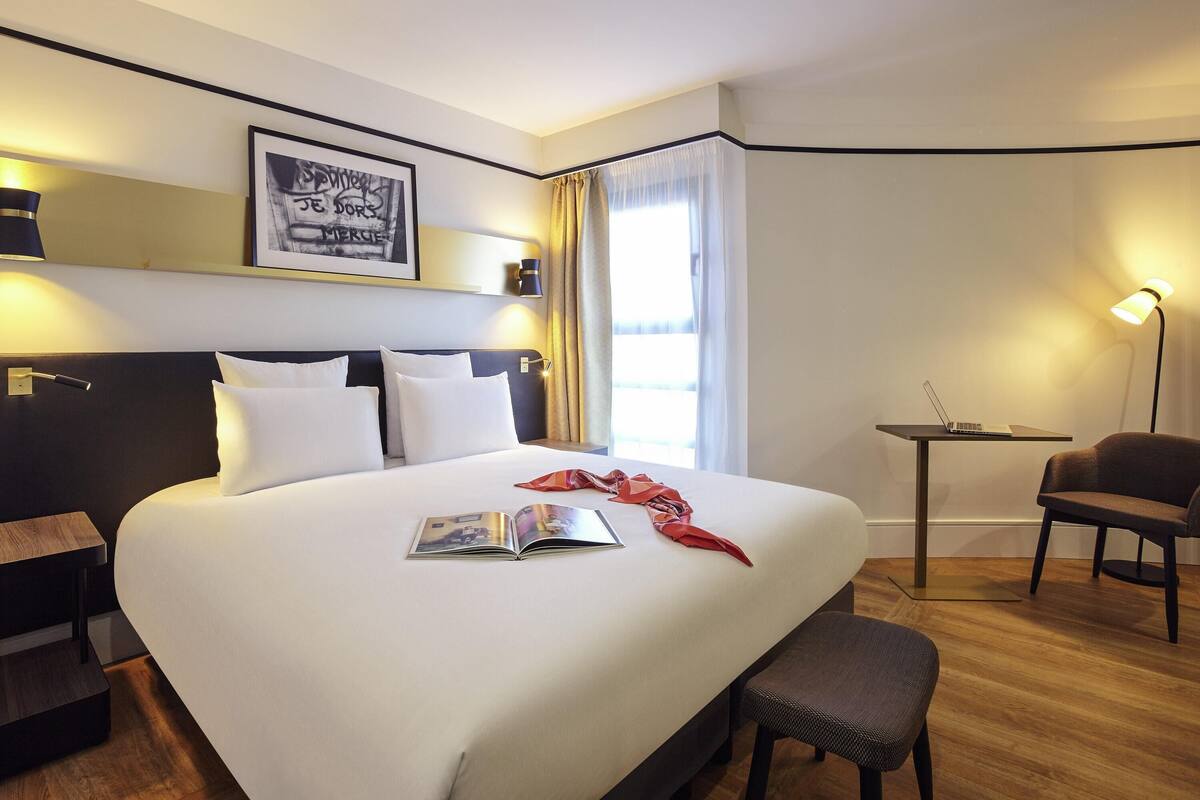 Quarto de hotel com uma cama grande, uma escrivaninha, uma cadeira, um abajur e um quadro na parede.