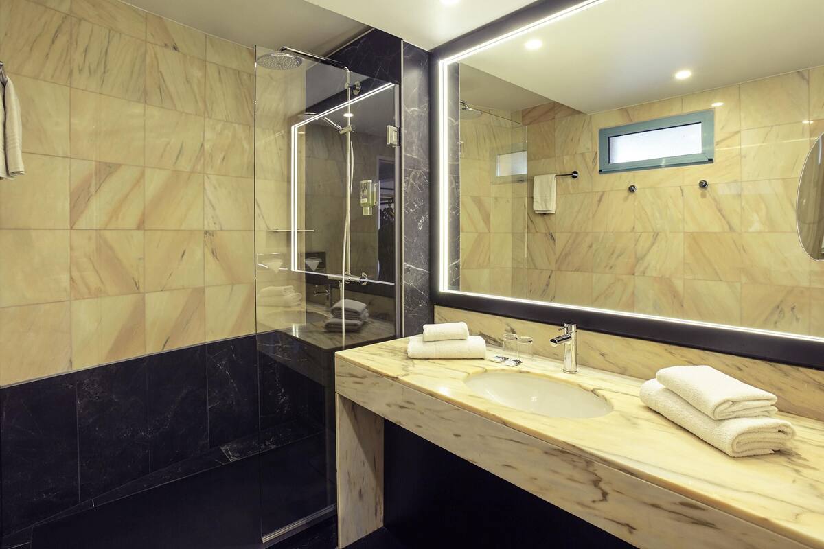 A modern bathroom with a large mirror, a glass-enclosed shower, and a marble-tiled wall.