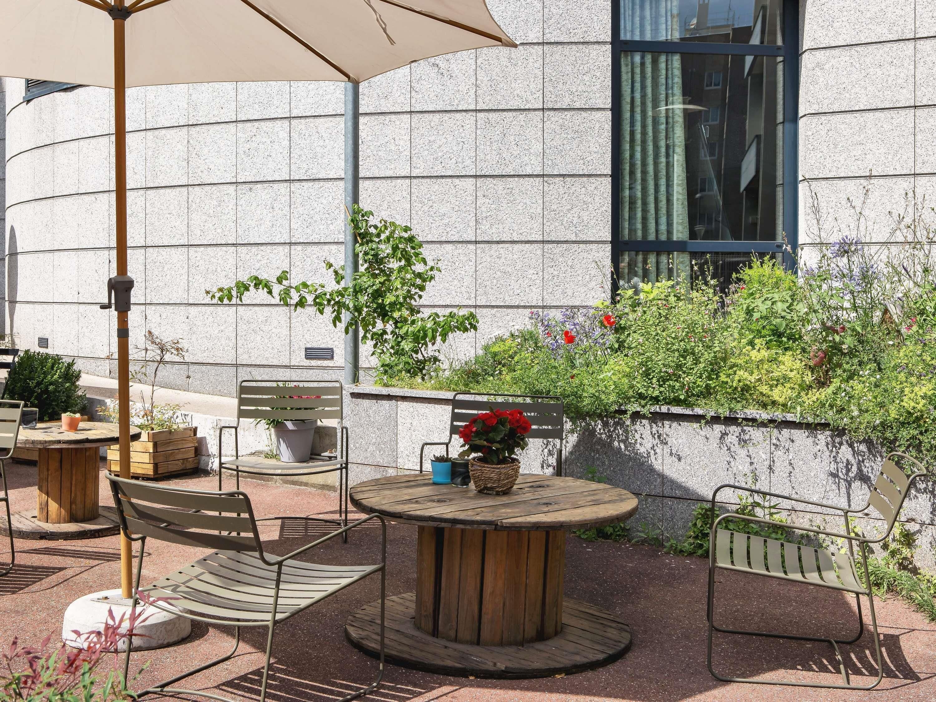 An outdoor seating area with round wooden tables and metal chairs, surrounded by potted plants and a stone wall.