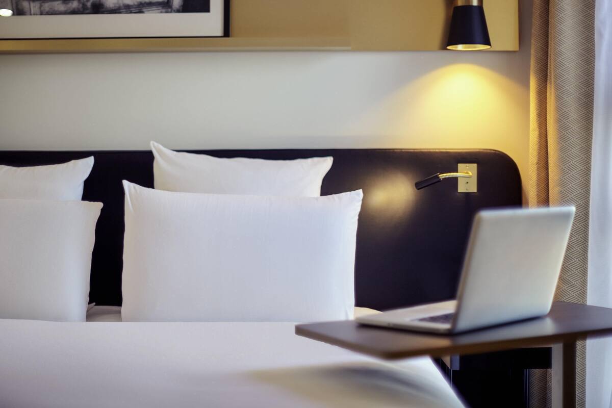 A hotel room with a bed, white pillows, a black headboard, a wall-mounted lamp, and a laptop on a small table.
