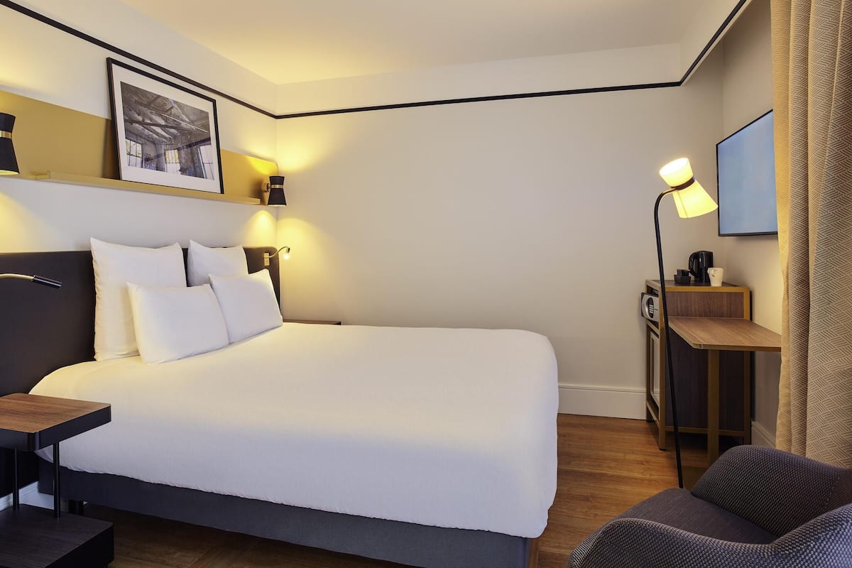 Quarto de hotel com uma cama grande, mesas de cabeceira, escrivaninha, cadeira, televisão e um quadro na parede.