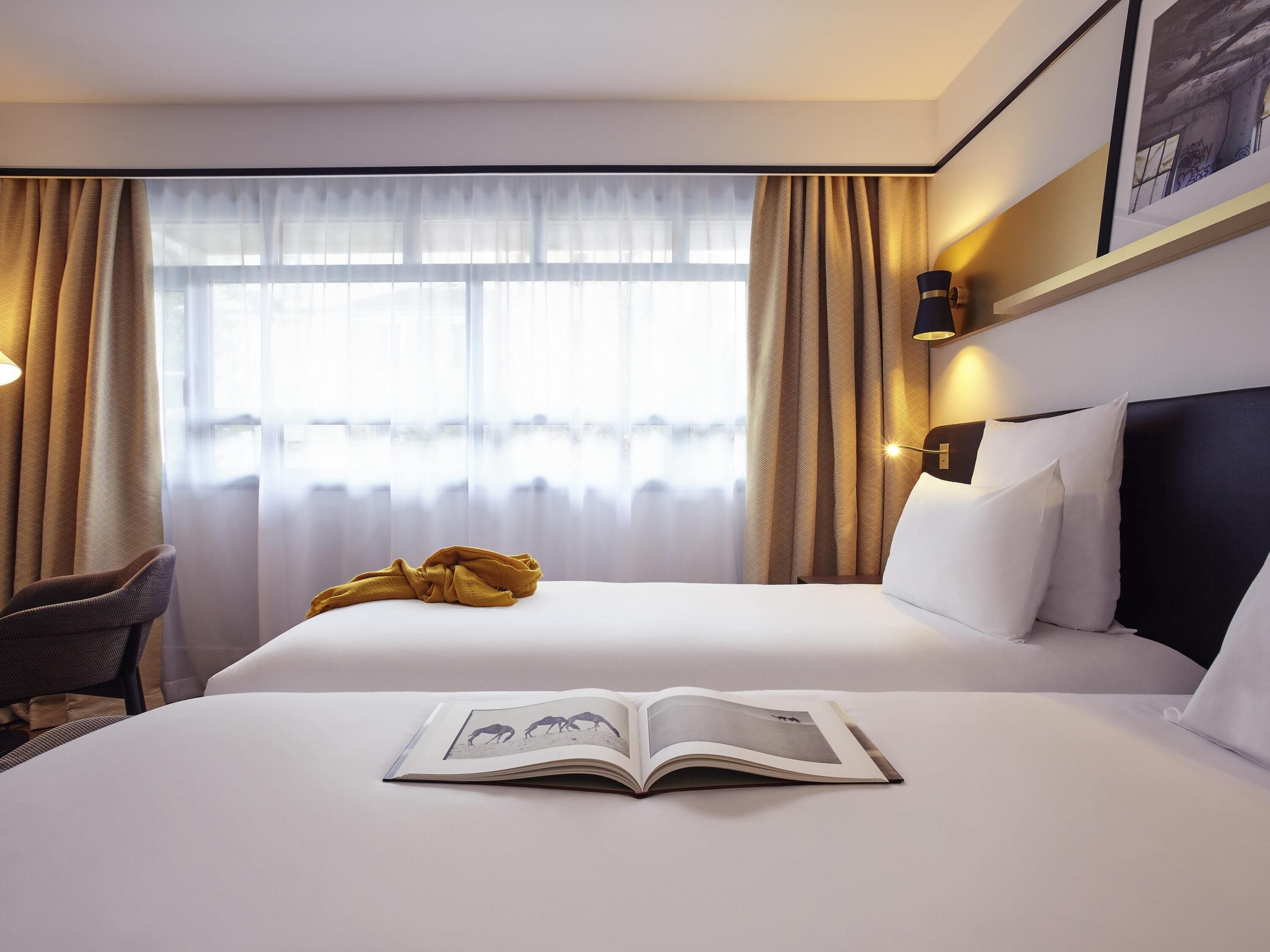 A hotel room with a bed, a chair, a book, and a window with curtains.