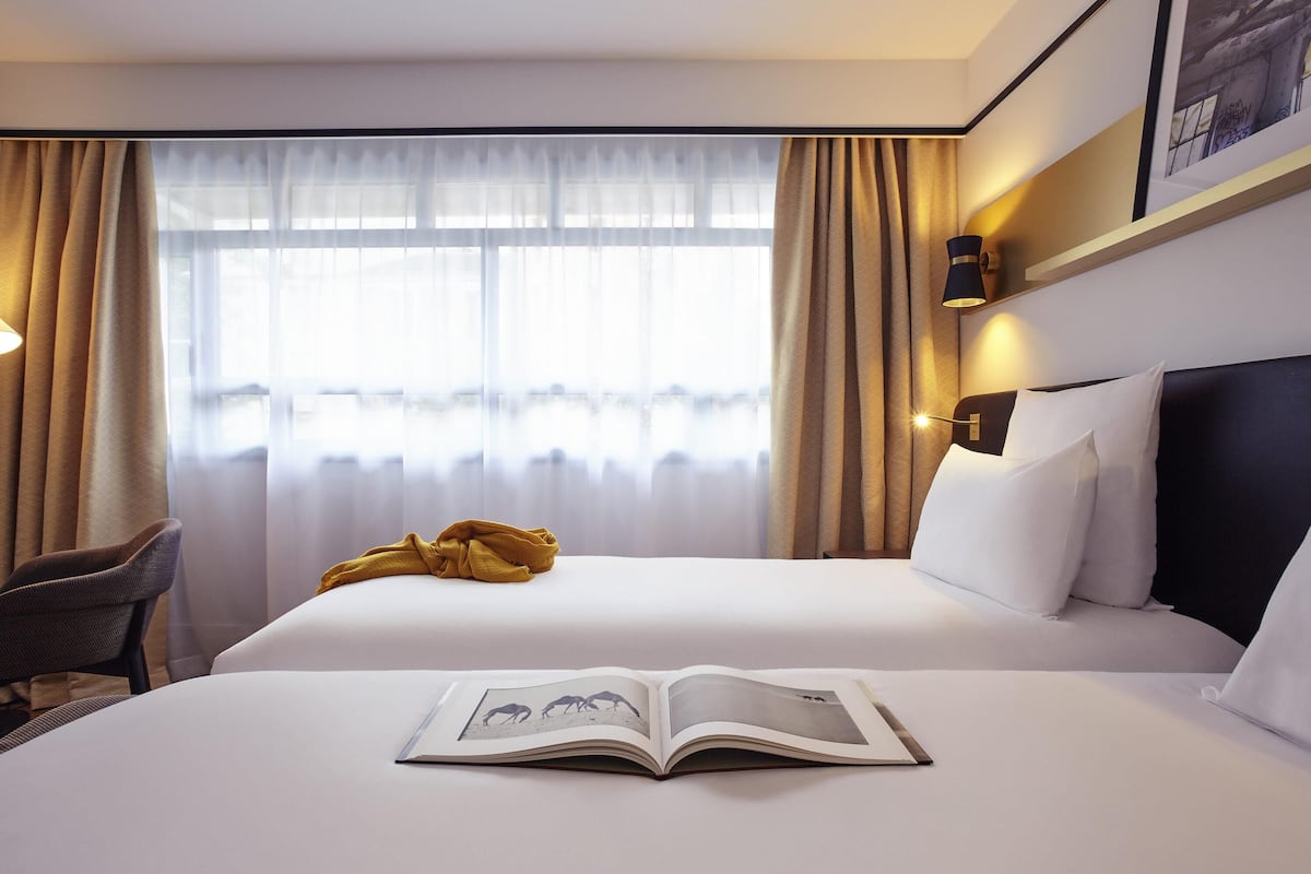 A hotel room with a bed, a chair, a book, and a window with curtains.