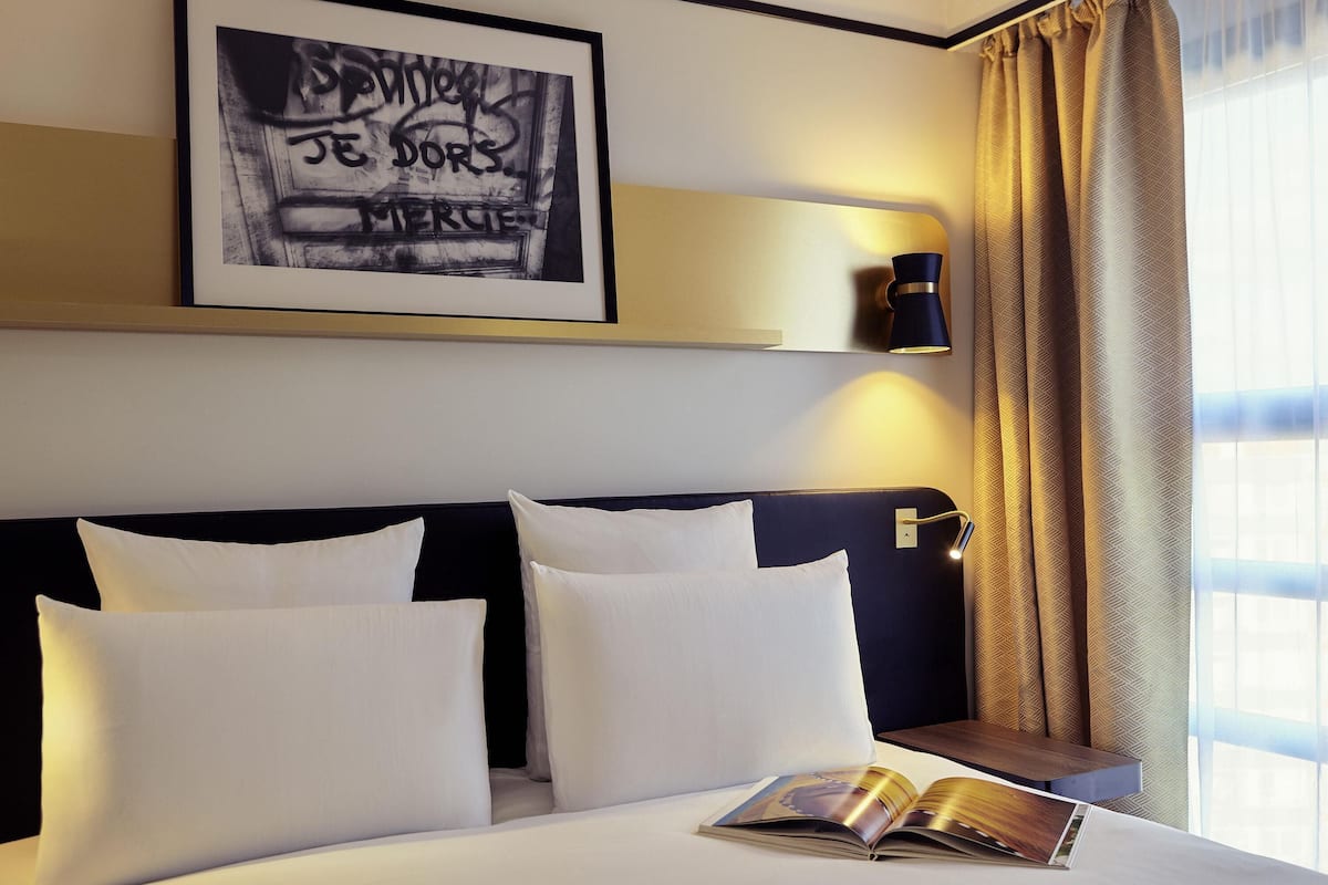 A hotel room with a bed, pillows, a framed artwork, a wall-mounted light, and a window with curtains.