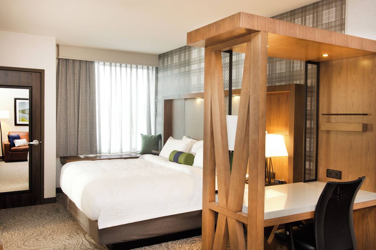 Habitación de hotel con una cama grande, un escritorio con silla, un espejo y una ventana con cortinas.