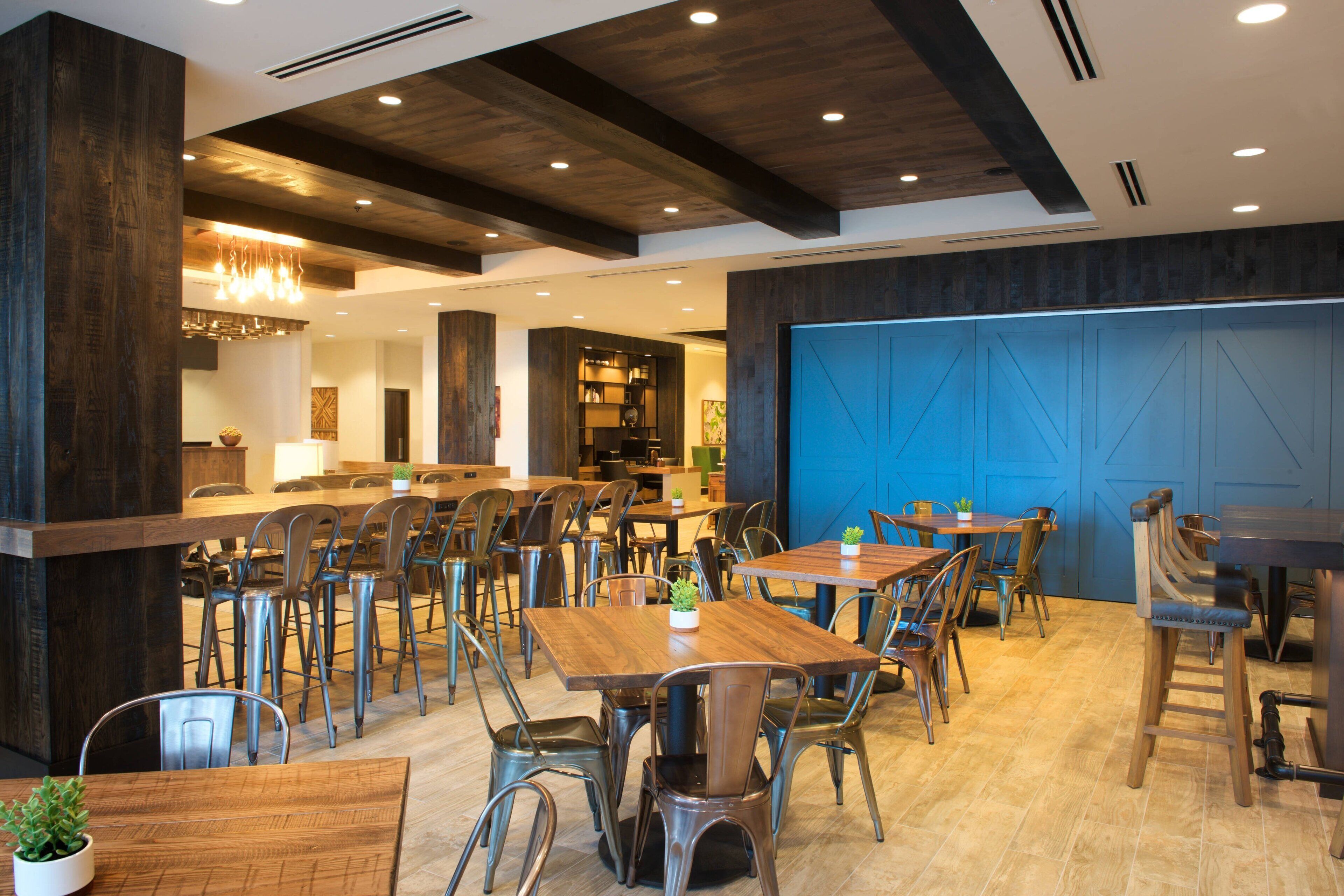 A modern restaurant with wooden tables and metal chairs, a blue accent wall, and a chandelier.