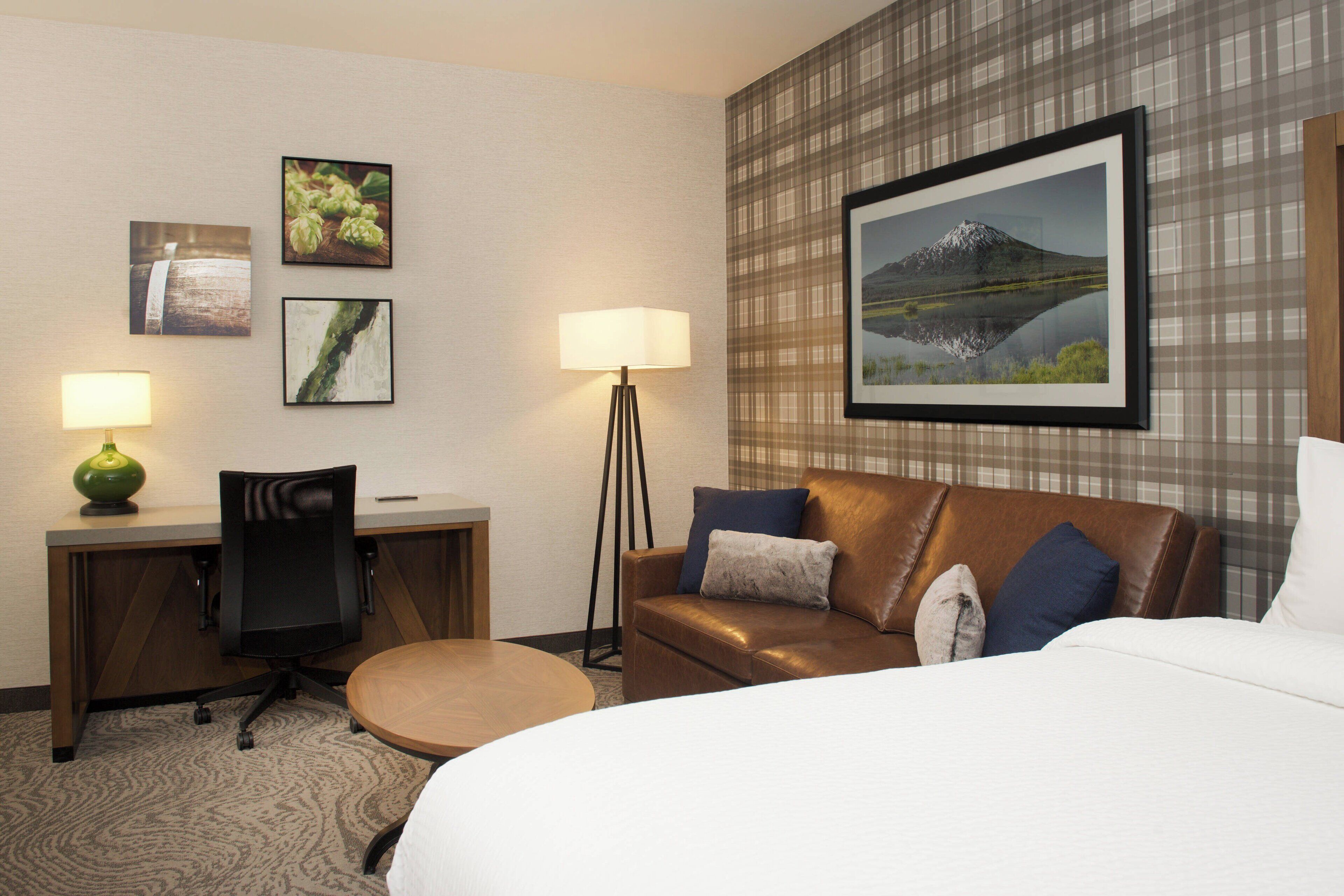 A hotel room with a bed, a desk with a chair, a sofa, and two lamps. There are framed pictures on the wall.