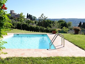 Une piscine rectangulaire avec un escalier en métal sur une terrasse carrelée, entourée de pelouse et de plantes en pot, et offrant une vue sur une colline et des bâtiments au loin.