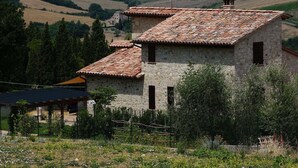 Un bâtiment en pierre, surmonté d’un toit en tuiles, entouré de verdure et de collines.