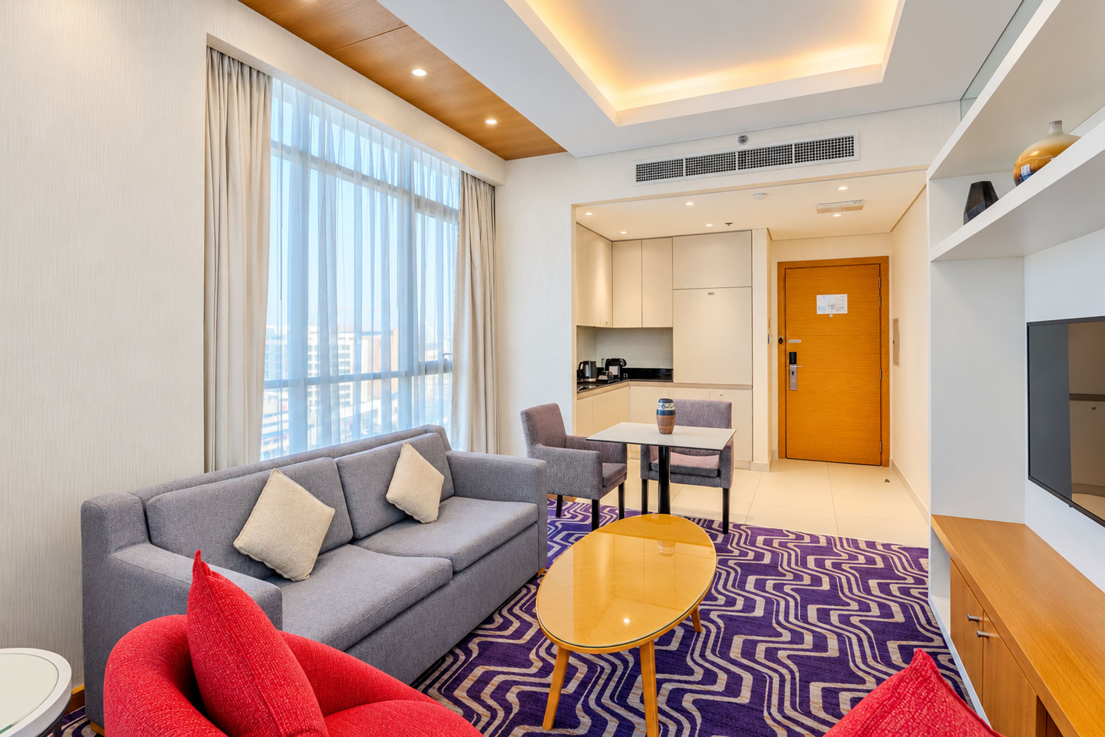 A modern hotel room with a sofa, a small table, a kitchen area, and a television.