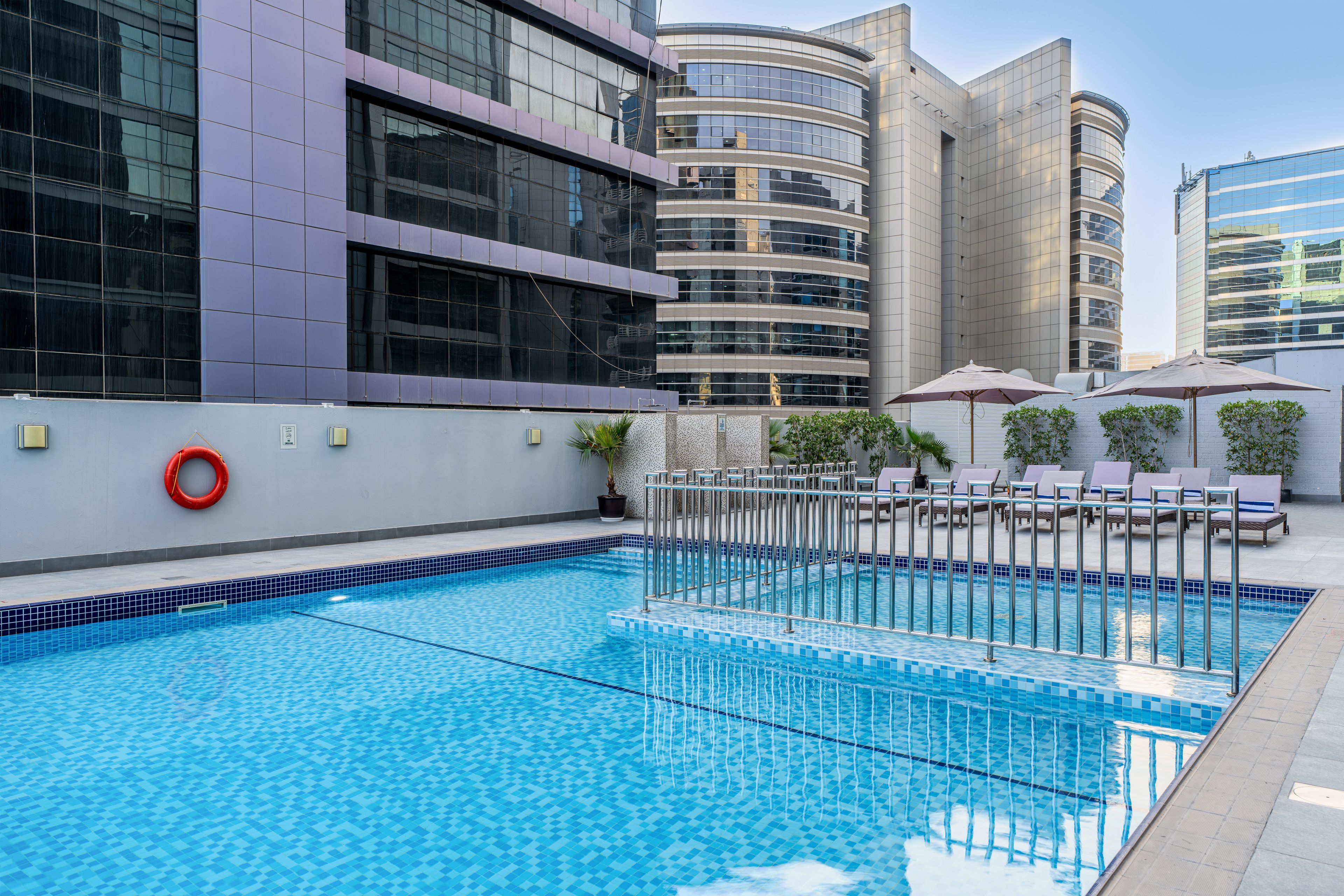 A swimming pool with a tiled edge, surrounded by lounge chairs and umbrellas, with modern buildings in the background.