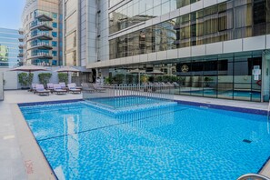 Une piscine dont le bord est revêtu de carreaux, entourée de chaises longues et de parasols, et située à côté d’un bâtiment moderne aux fenêtres en verre réfléchissant.