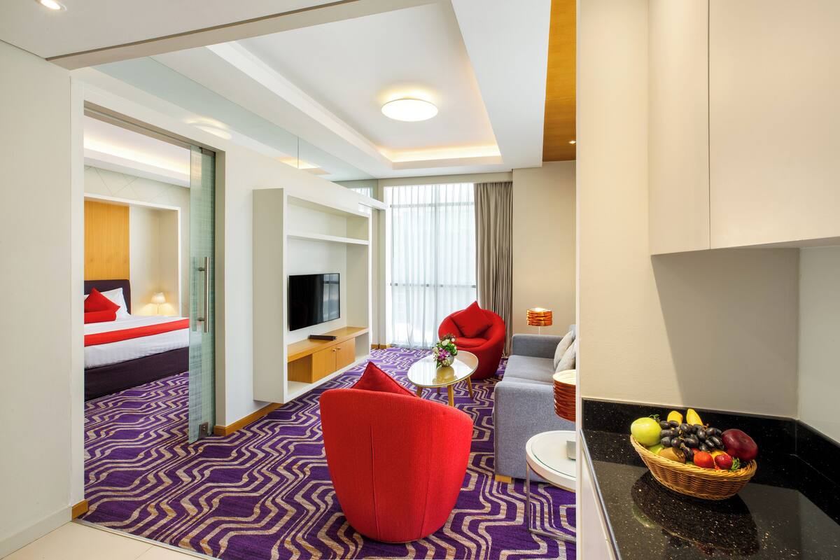 Une chambre d’hôtel avec un lit, un fauteuil rouge, un canapé, une télévision, une table à manger et un panier de fruits.