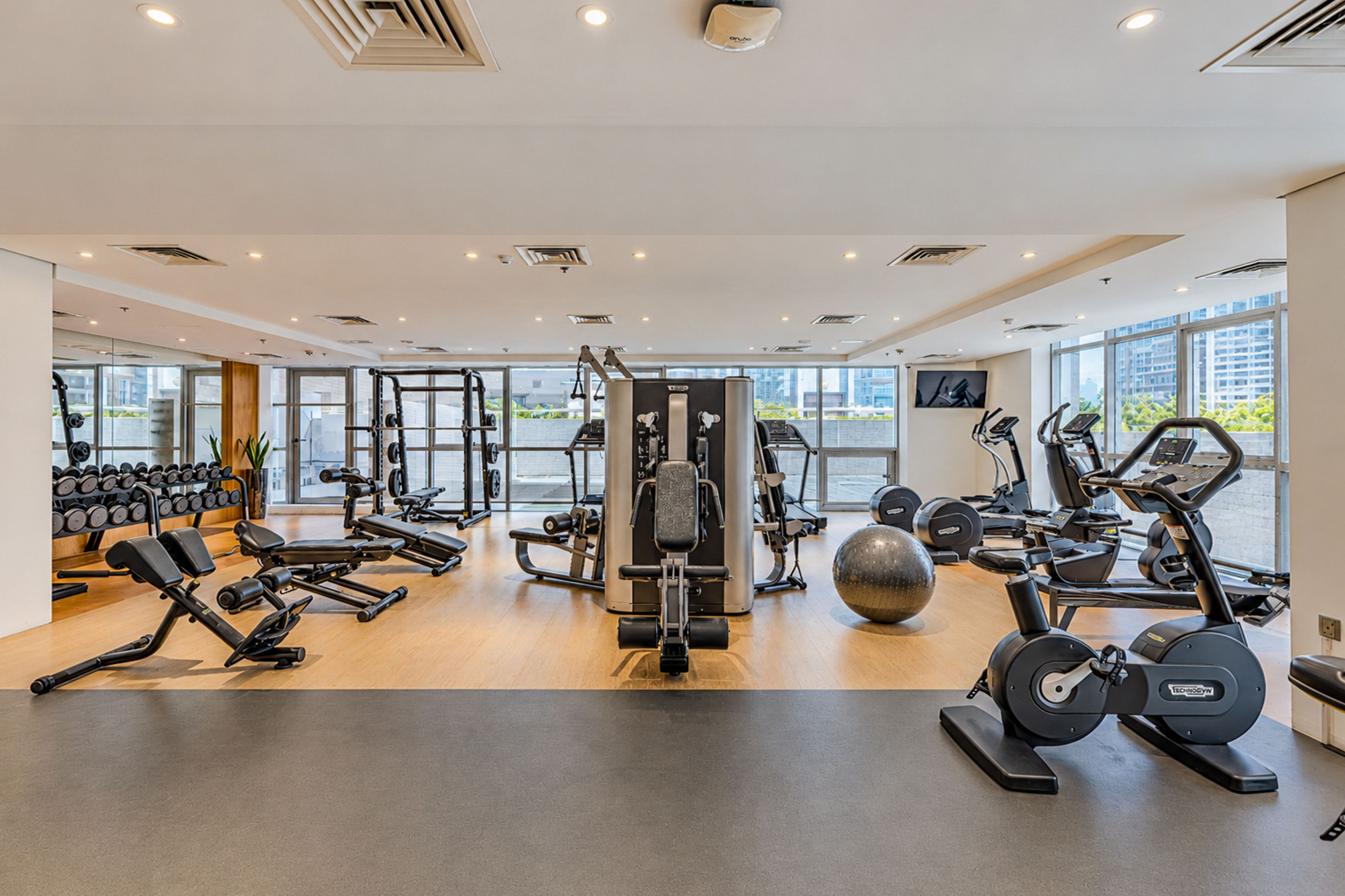 A modern gym with various exercise equipment, large windows, and a clean, organized layout.