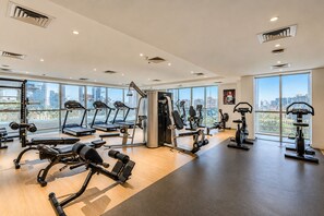Une salle de sport équipée de tapis de course, d’appareils de musculation et offrant une vue sur la ville grâce à de grandes fenêtres.