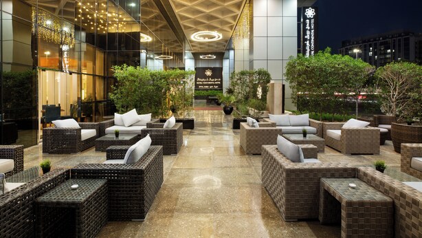 A modern hotel lobby with wicker furniture, a marble floor, and a sign indicating the hotel name. - Royal Continental Hotel (Dubai)