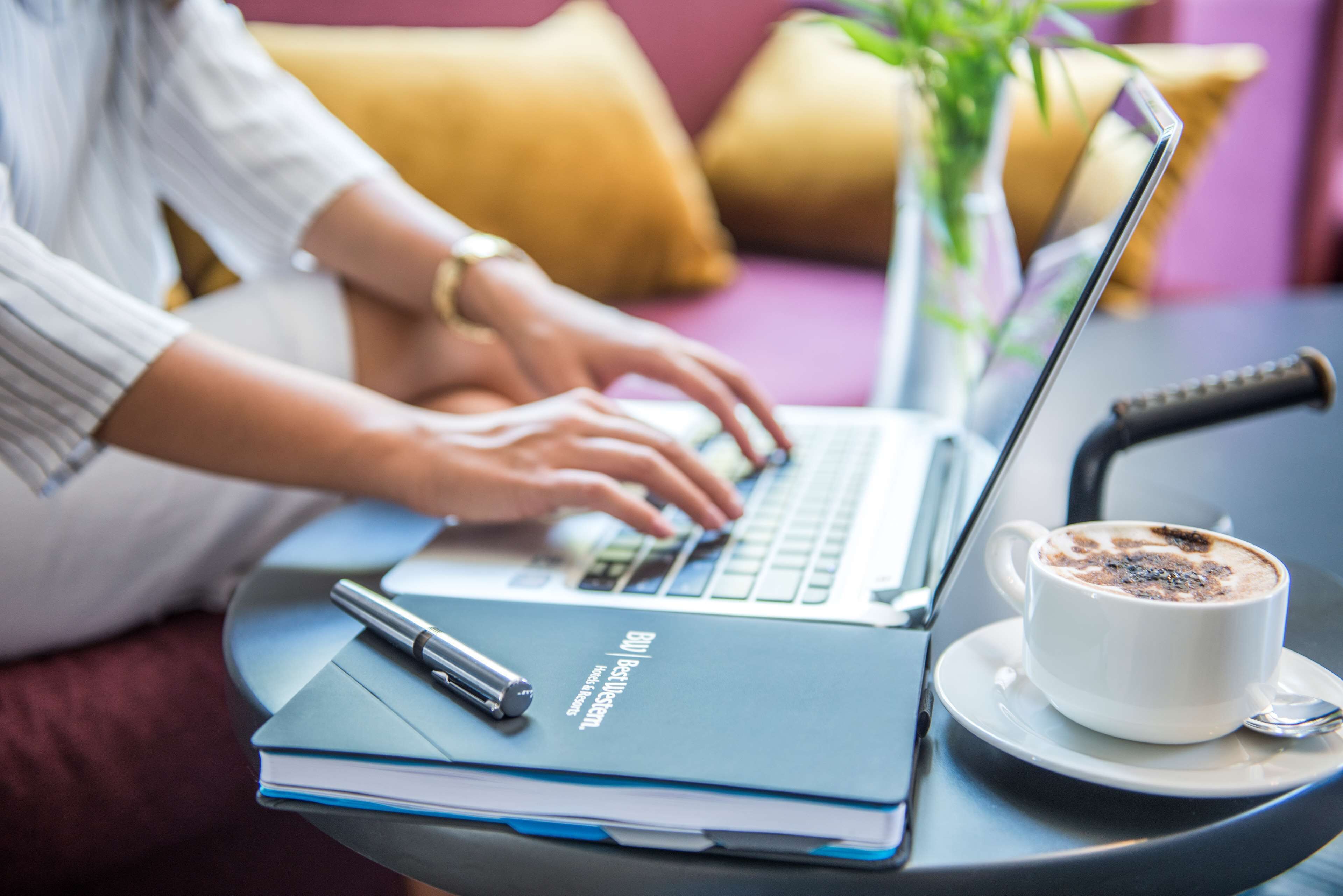 Una persona usando una computadora portátil sobre una mesa, con un cuaderno, un bolígrafo y una taza de café.