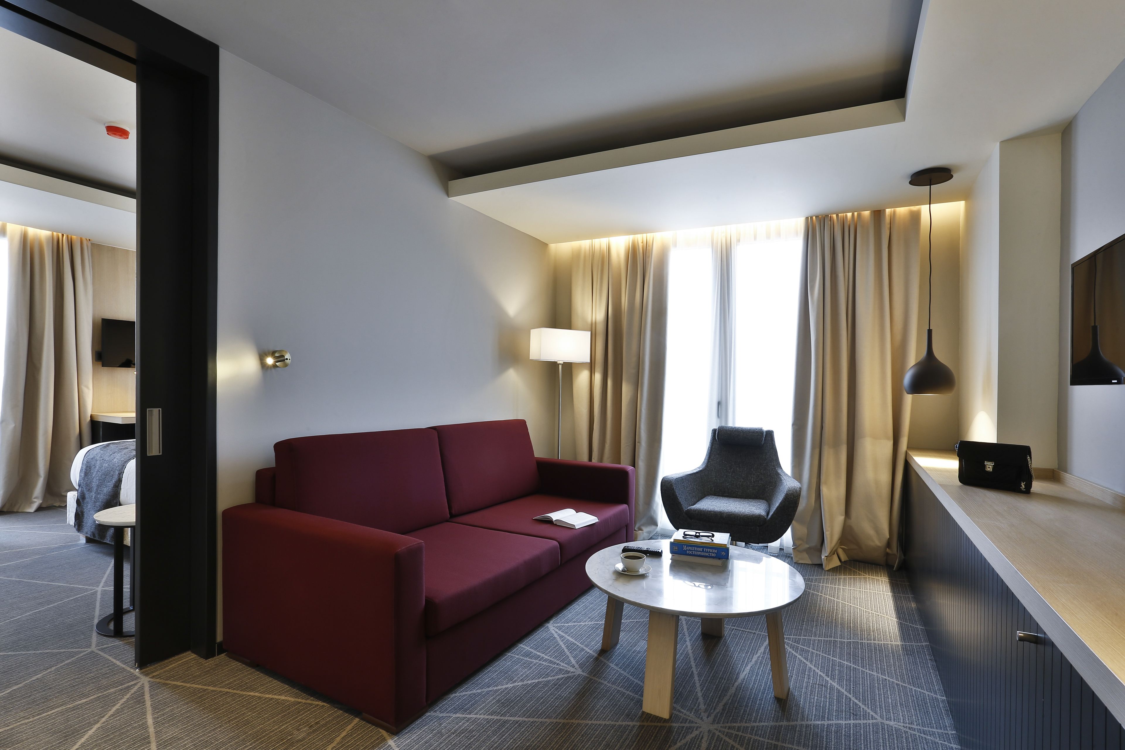 Habitación de hotel con sofá rojo, una pequeña mesa redonda, una silla, una lámpara y vistas al exterior a través de amplios ventanales con cortinas.
