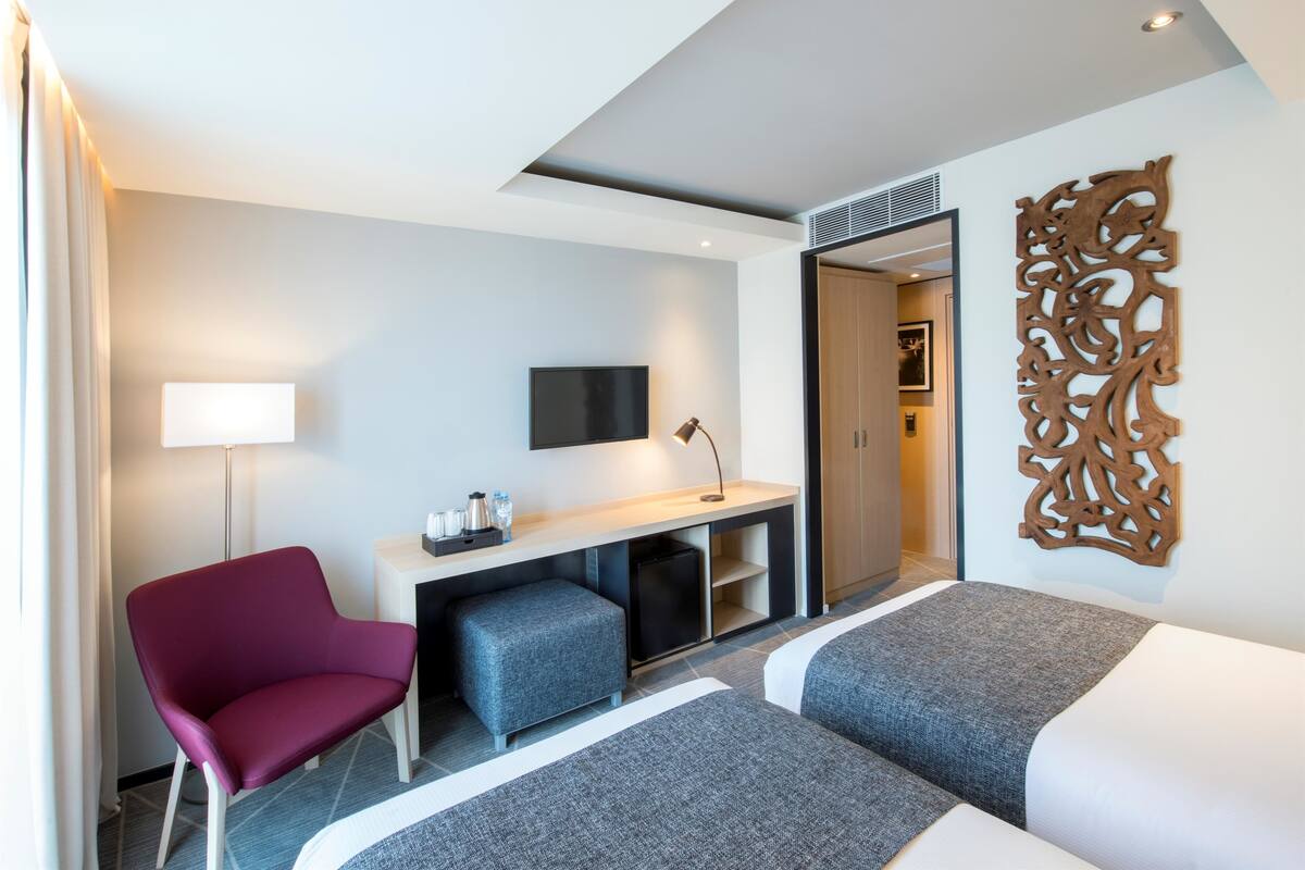 A hotel room with a desk, two beds, a chair, a TV, and a decorative wall piece.