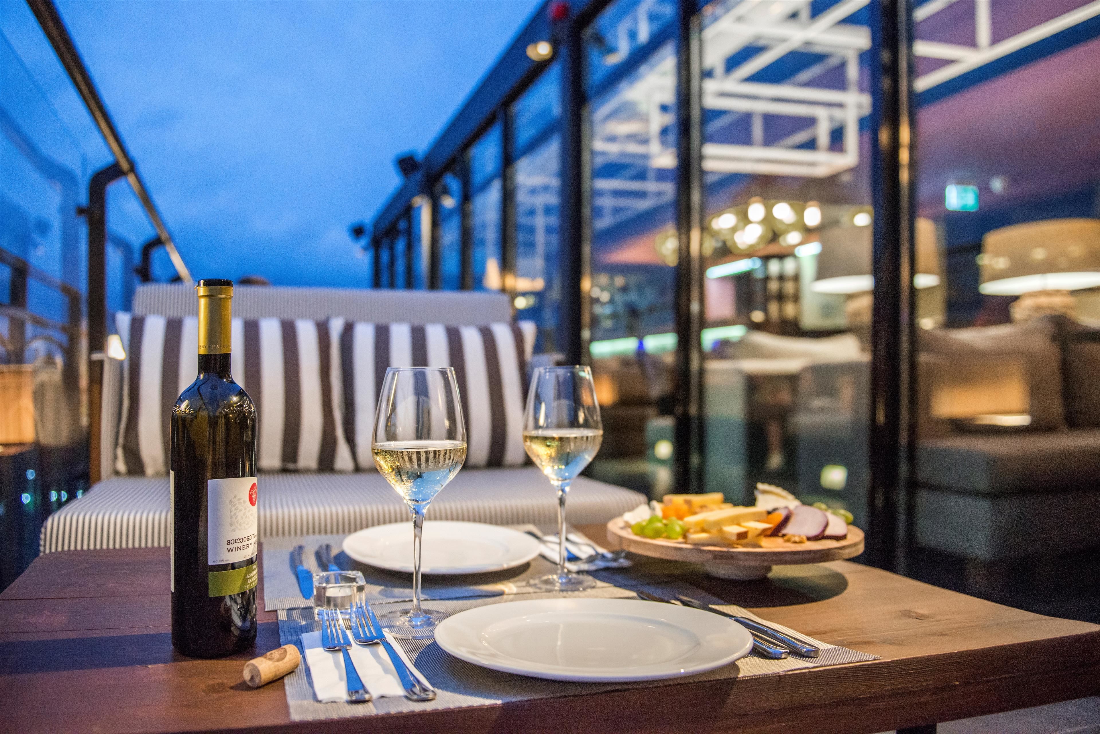 A table set with a bottle of wine, two glasses of white wine, plates, cutlery, and a small dish of food on a balcony with a view of the building's interior.