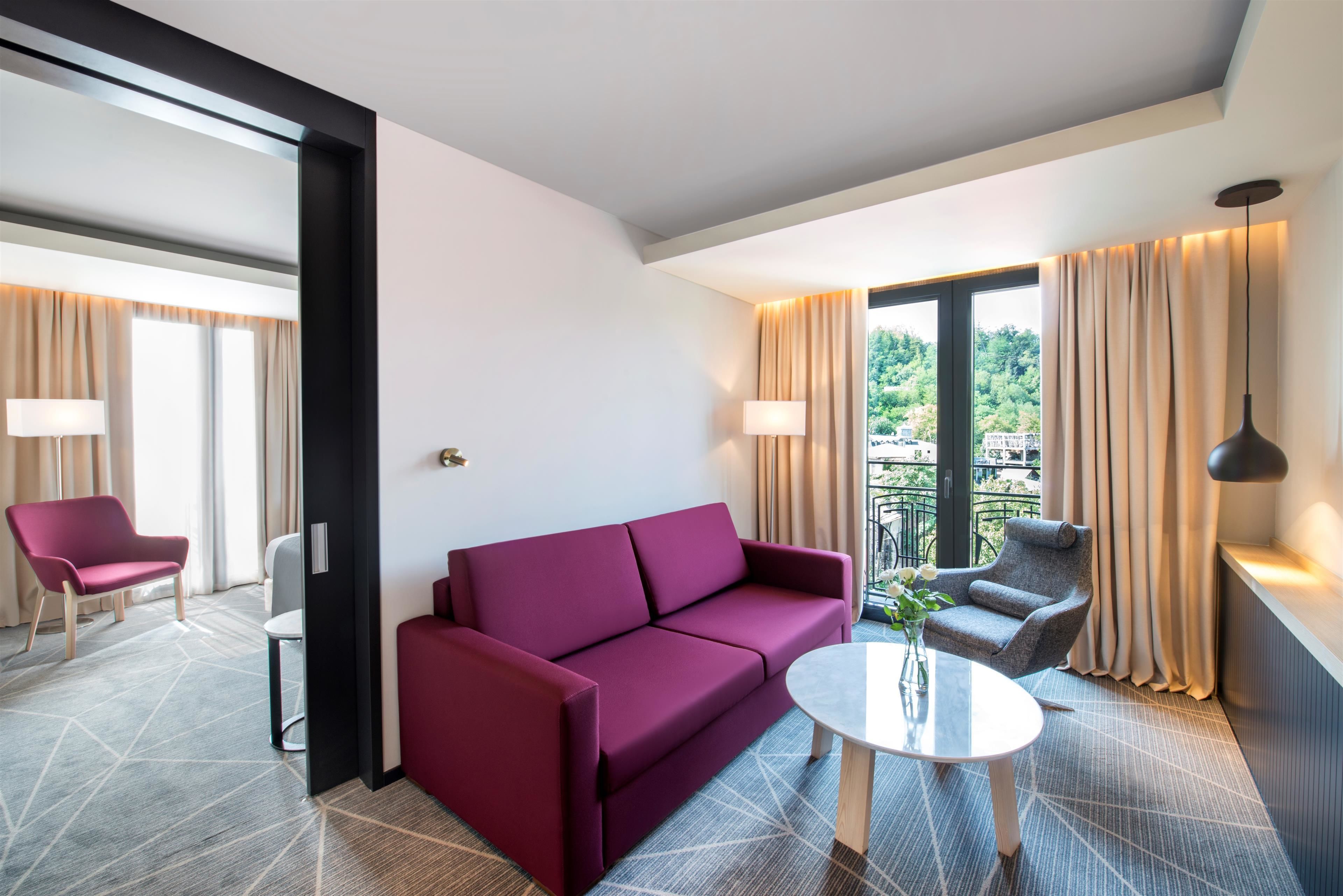 A modern hotel room with a red sofa, a grey armchair, a round table, and a balcony with curtains.