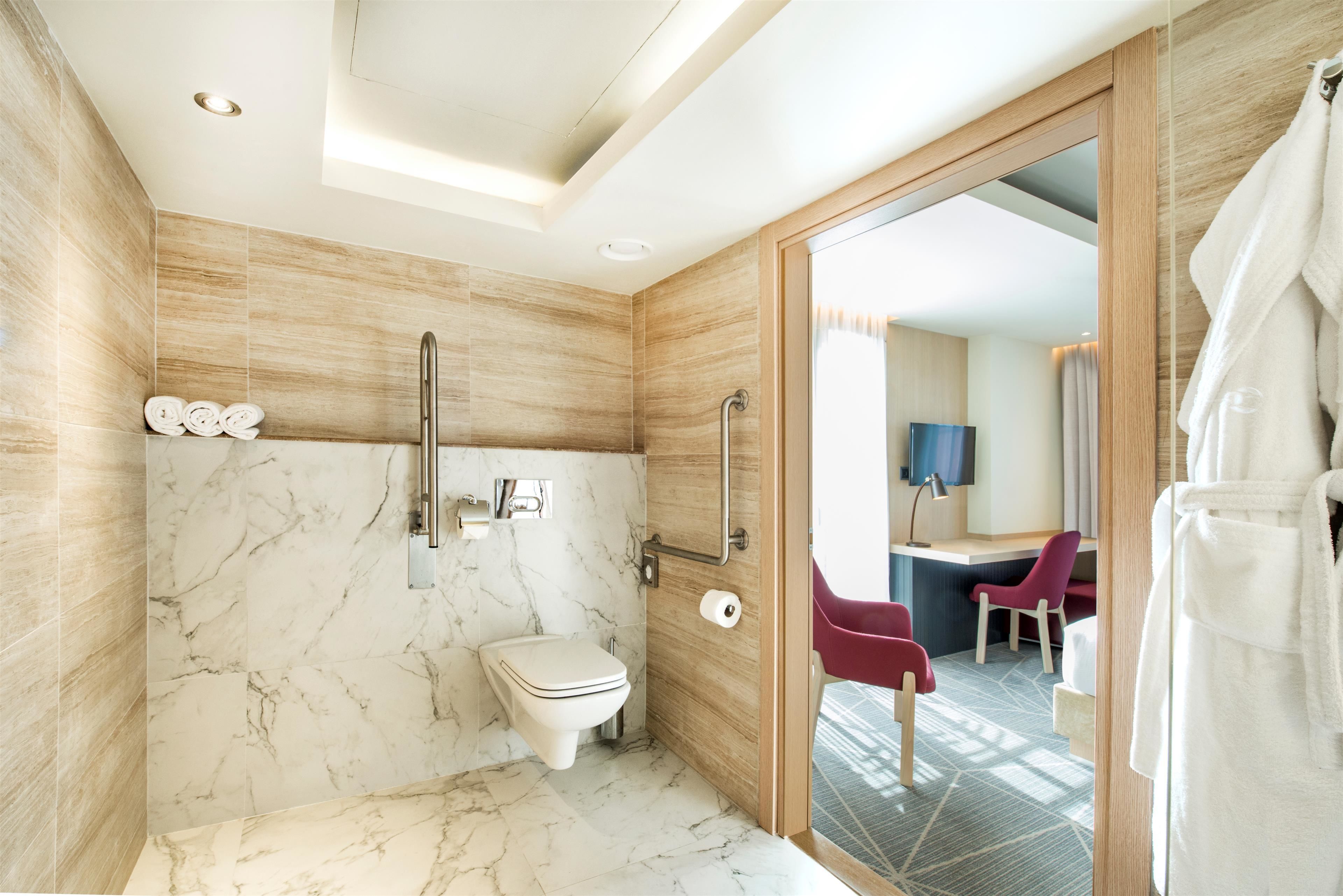 Une salle de bain moderne avec un sol en marbre, des toilettes, une douche et une porte en verre donnant sur une pièce équipée d’un bureau et de chaises.