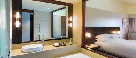 A hotel bathroom with a large mirror, a sink, a bathtub, and a bed with a headboard.