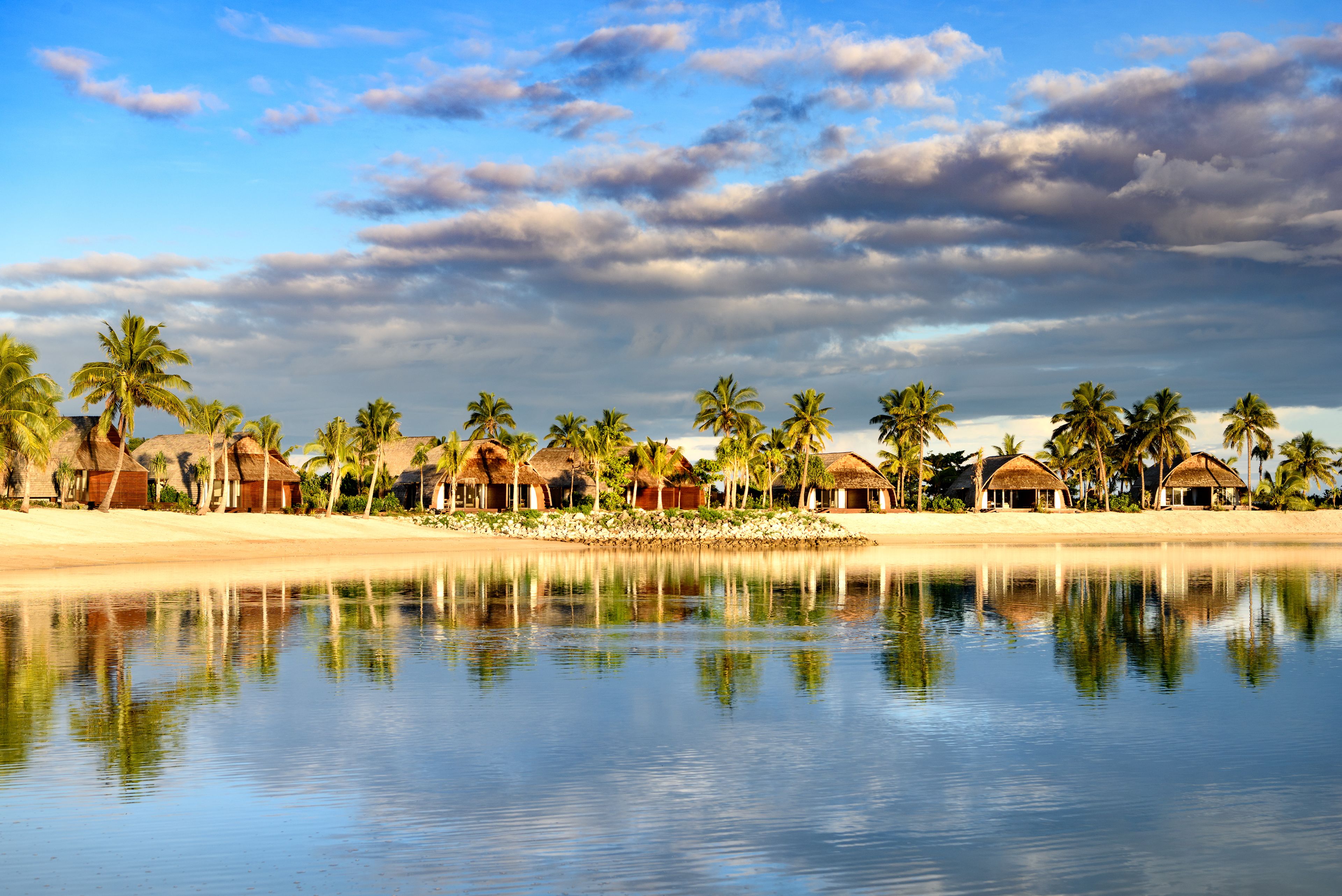 Un resort frente a la playa con cabañas de techo de paja, palmeras y un lago tranquilo que refleja el cielo.
