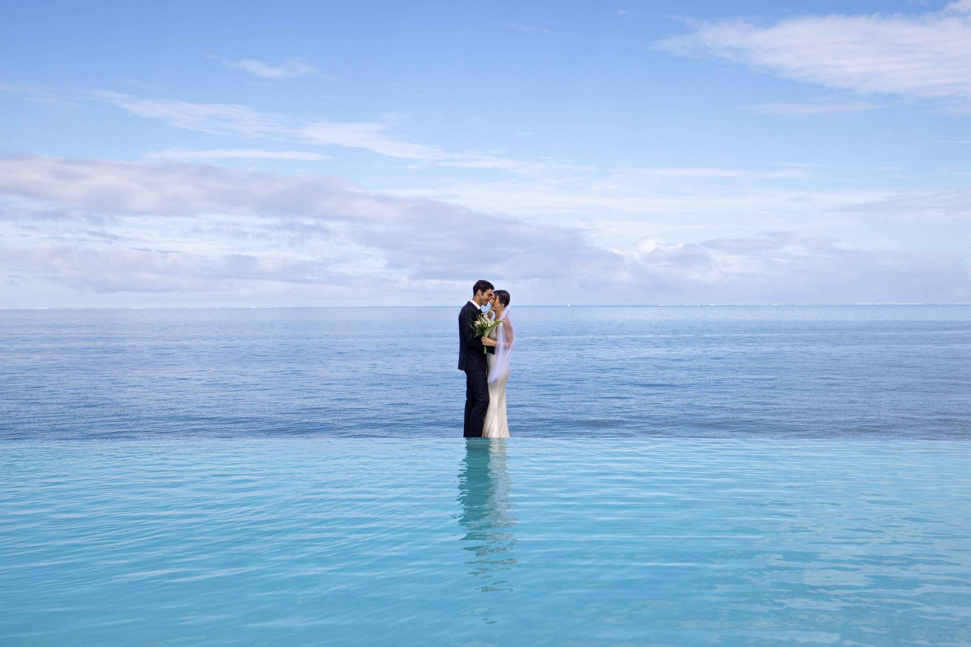 A couple standing in a clear blue pool with a view of the ocean.