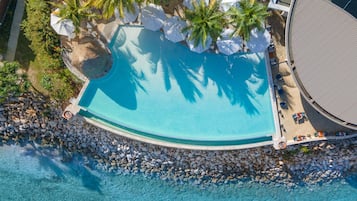 Vista aérea de un complejo turístico con piscina rodeada de palmeras y sombrillas, una zona de playa con rocas y un área de descanso cubierta.