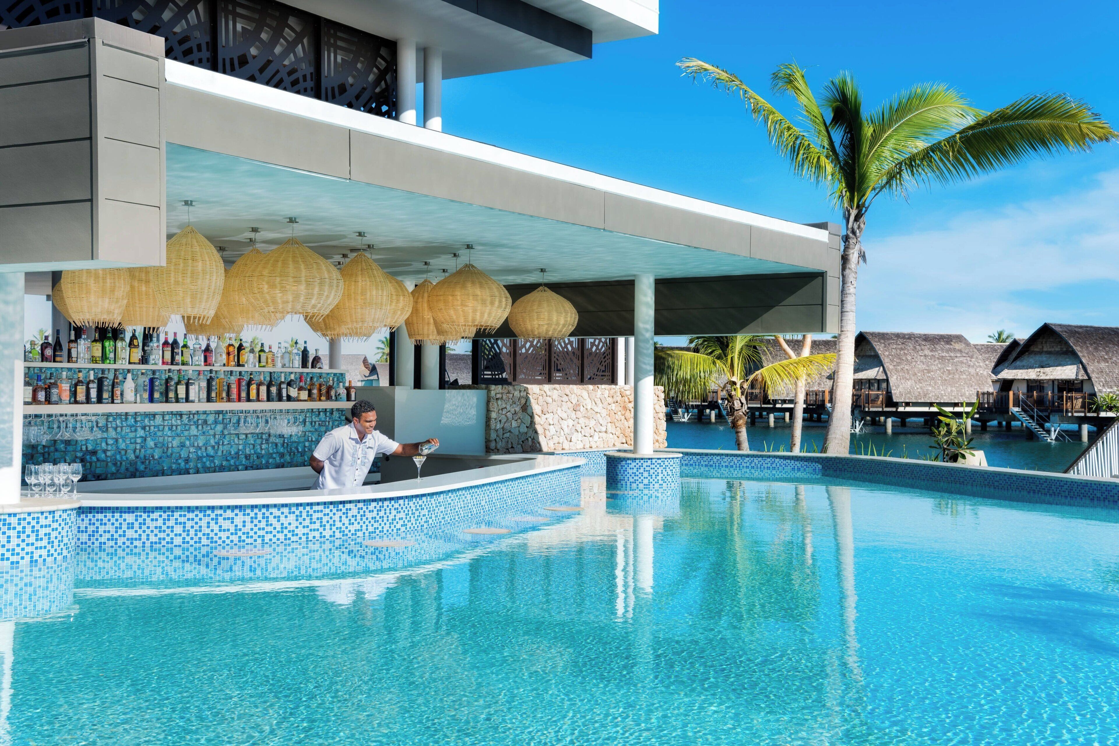 Uma área de piscina moderna com bar, espreguiçadeiras e bangalôs sobre a água.