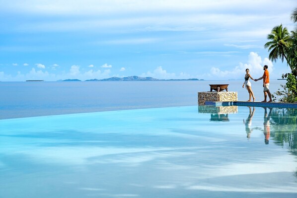 A couple standing by a pool with a view of the ocean and palm trees.