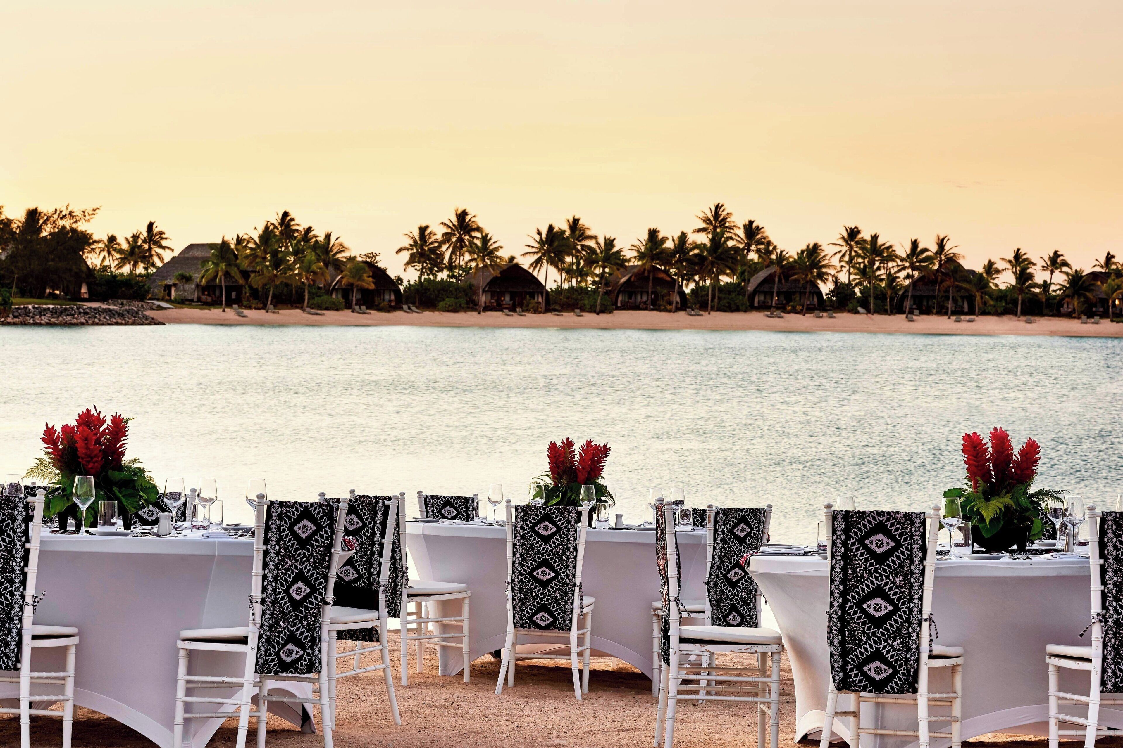 A beachfront dining setup with white tables and chairs, decorated with patterned tablecloths and black chair covers, overlooking a sandy beach with palm trees and huts.