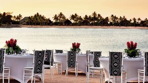 A beachfront dining setup with white tables and chairs, decorated with patterned tablecloths and black chair covers, overlooking a sandy beach with palm trees and huts.