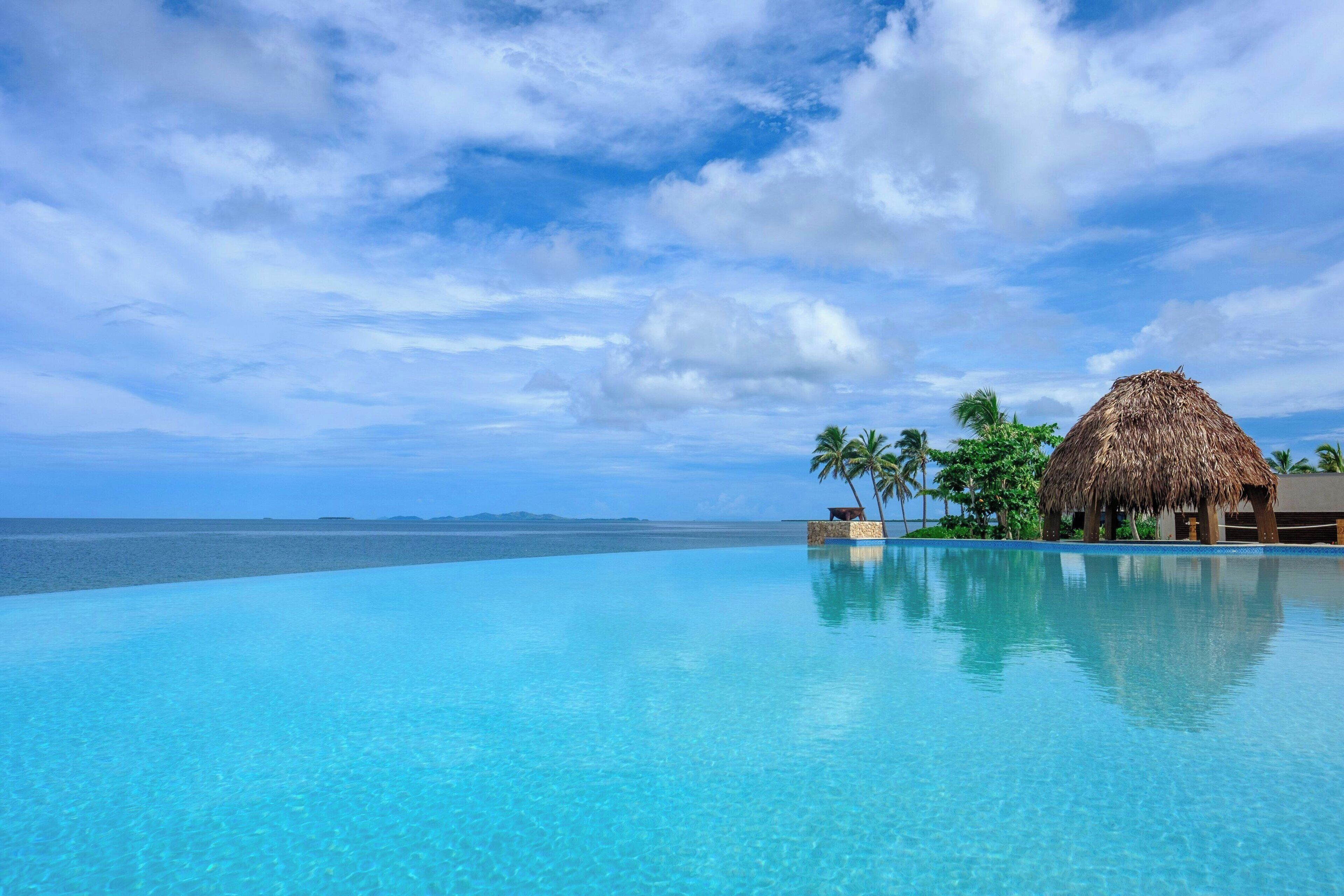 Una piscina infinita azul cristalina con una estructura de techo de paja y palmeras al fondo.