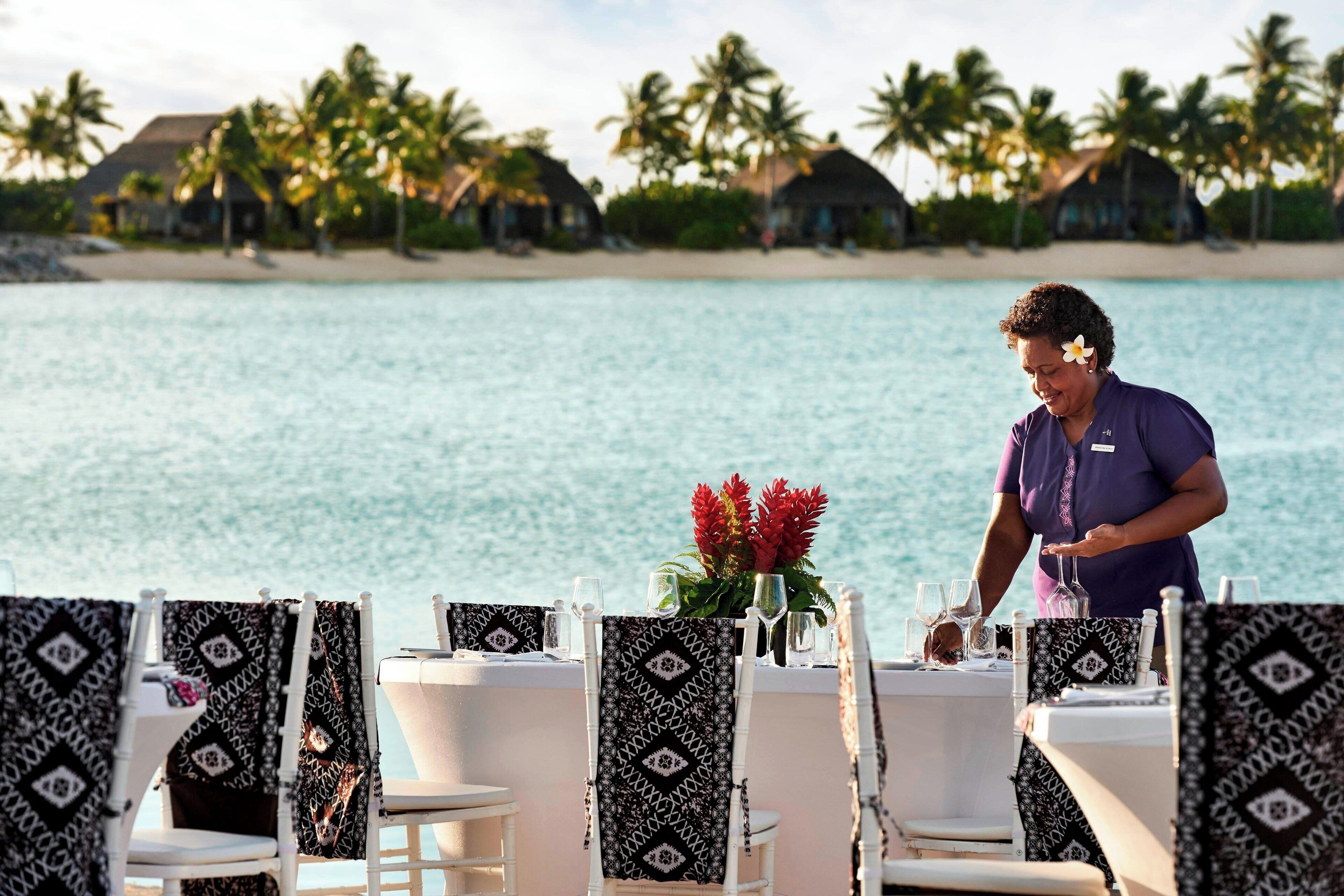 A beachside dining area with white chairs and black patterned tablecloths, overlooking a calm sea and palm trees.