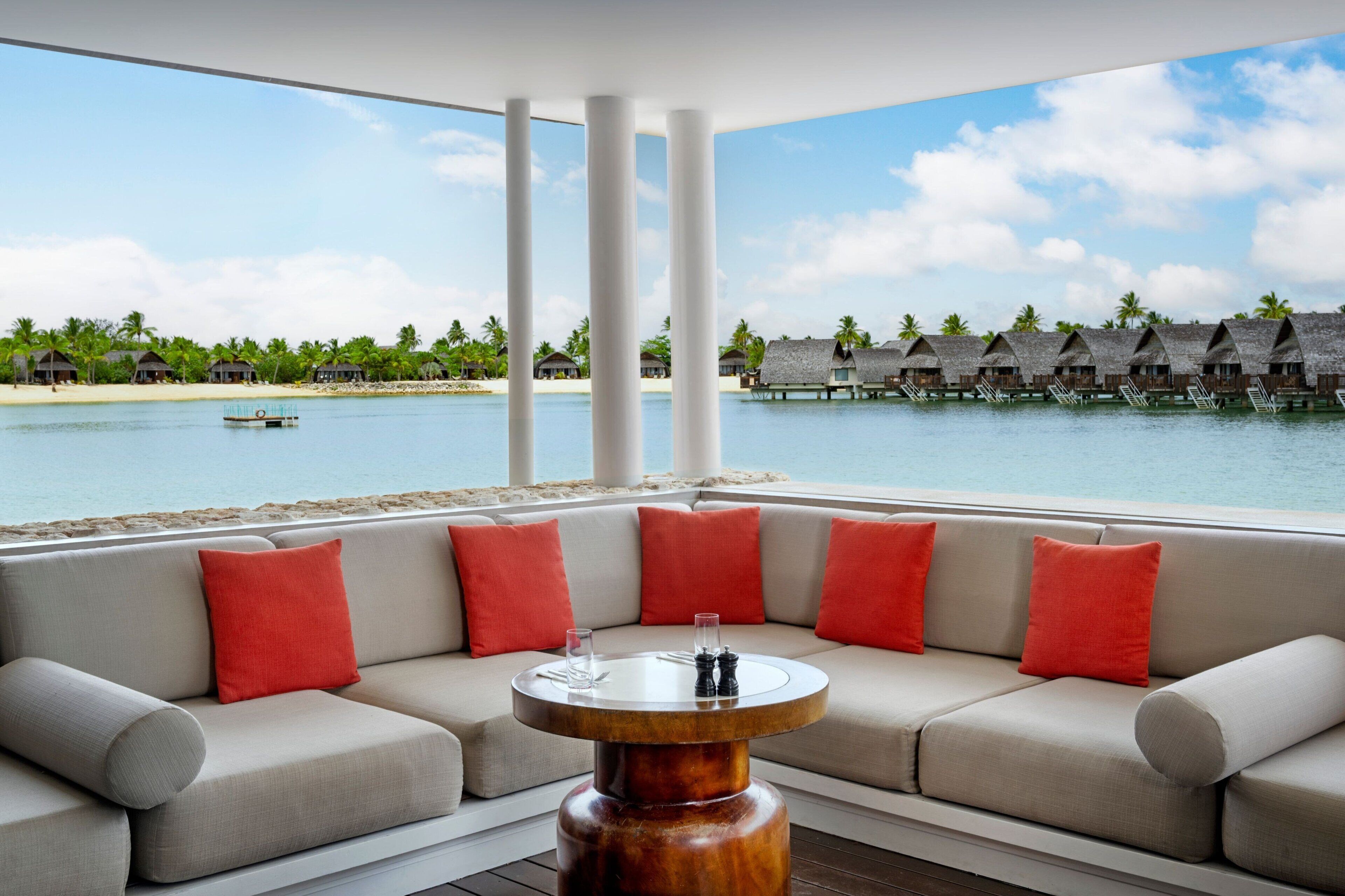 A modern lounge with a sofa and a round wooden table, overlooking a beach with overwater bungalows.