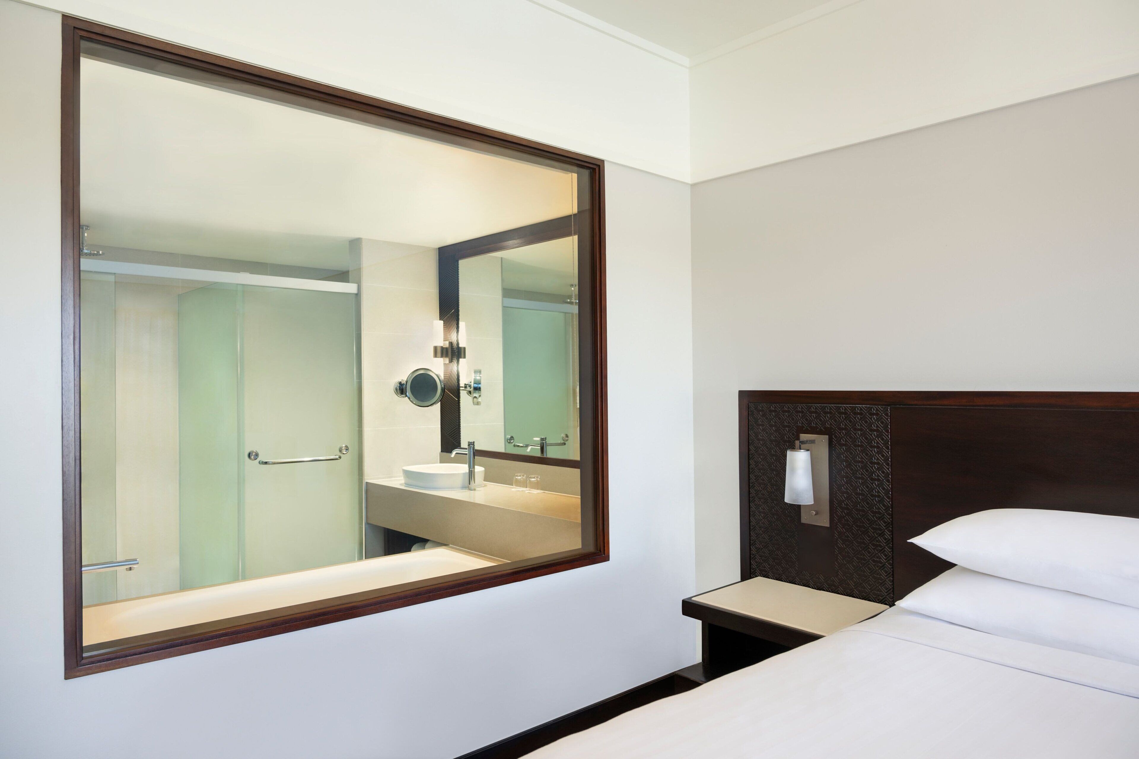 Habitación de hotel con un espejo grande, cama con ropa de cama blanca, mesita de noche y baño con lavabo y espejo.