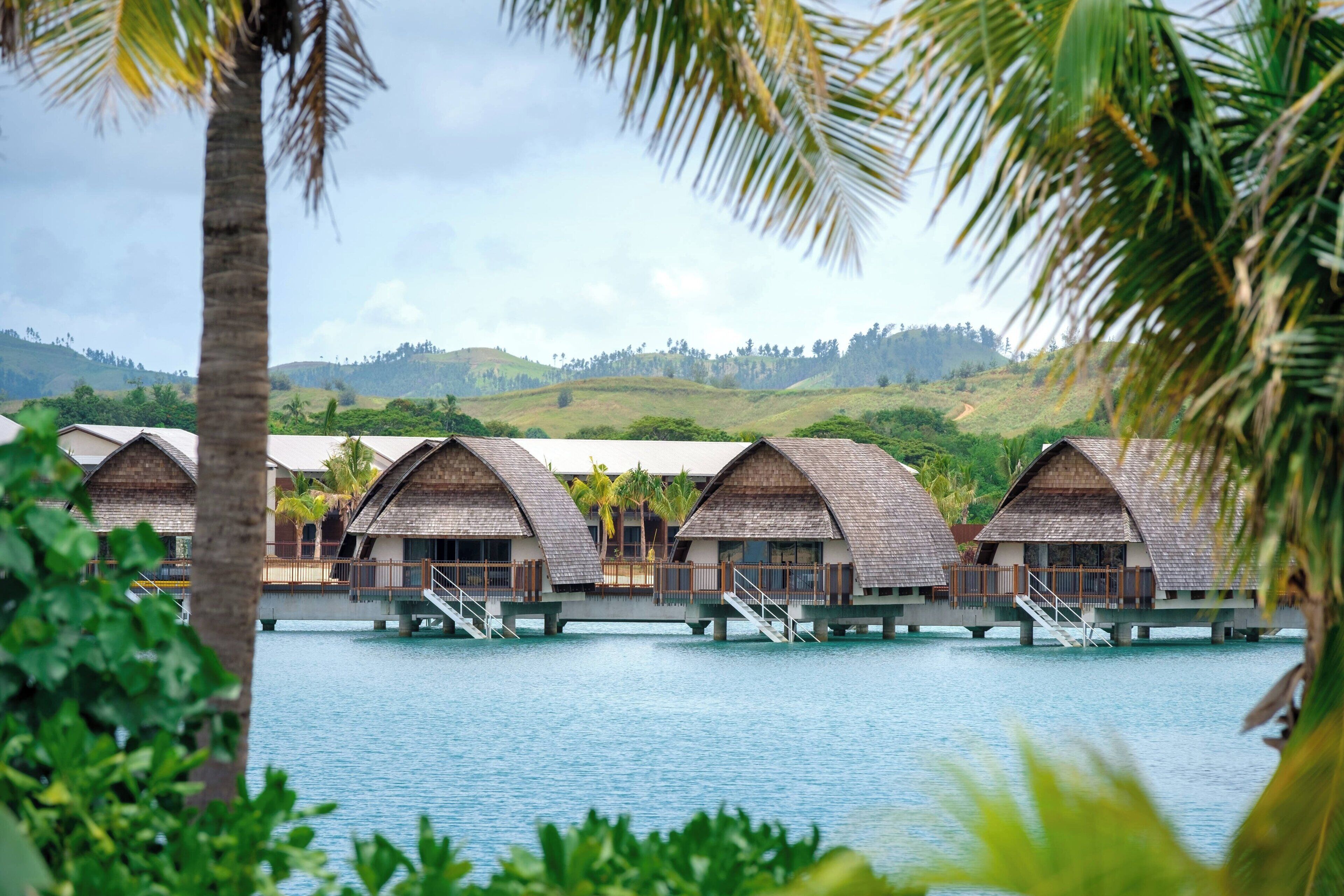 Wasserbungalows mit Holzdächern, verbunden durch Wege über ruhigem Wasser, umgeben von üppiger Vegetation und Hügeln.