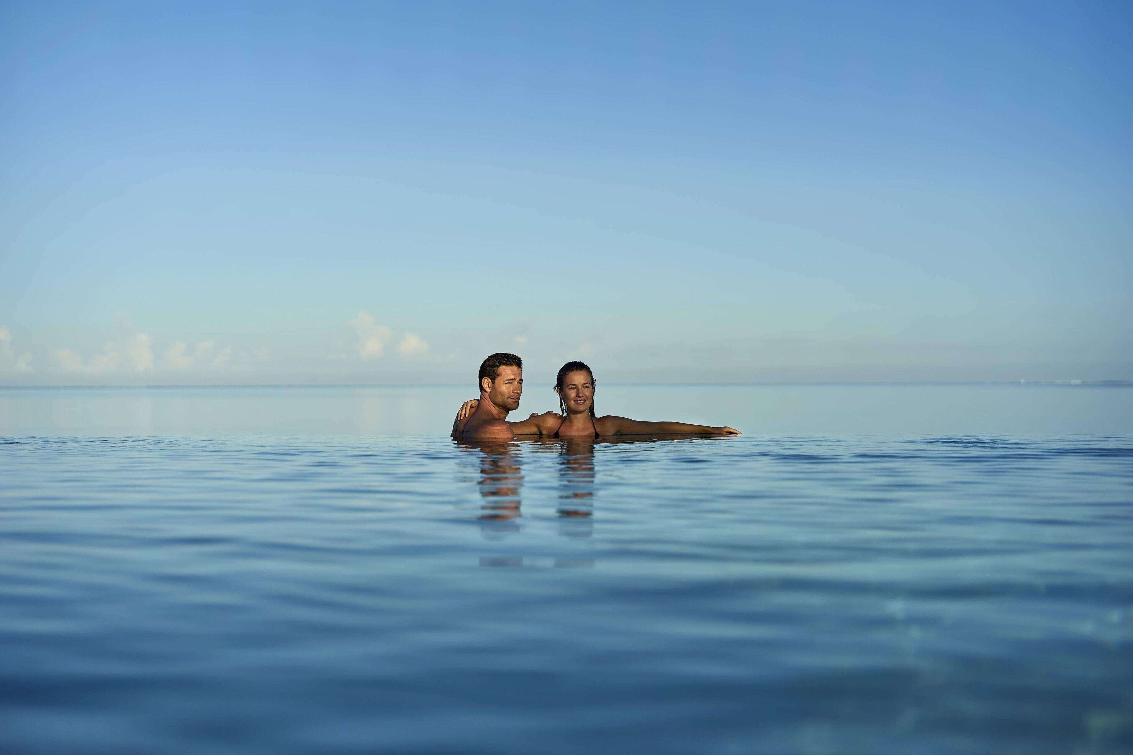 Dos personas en una amplia extensión de agua, posiblemente una piscina o laguna, con cielo despejado al fondo.