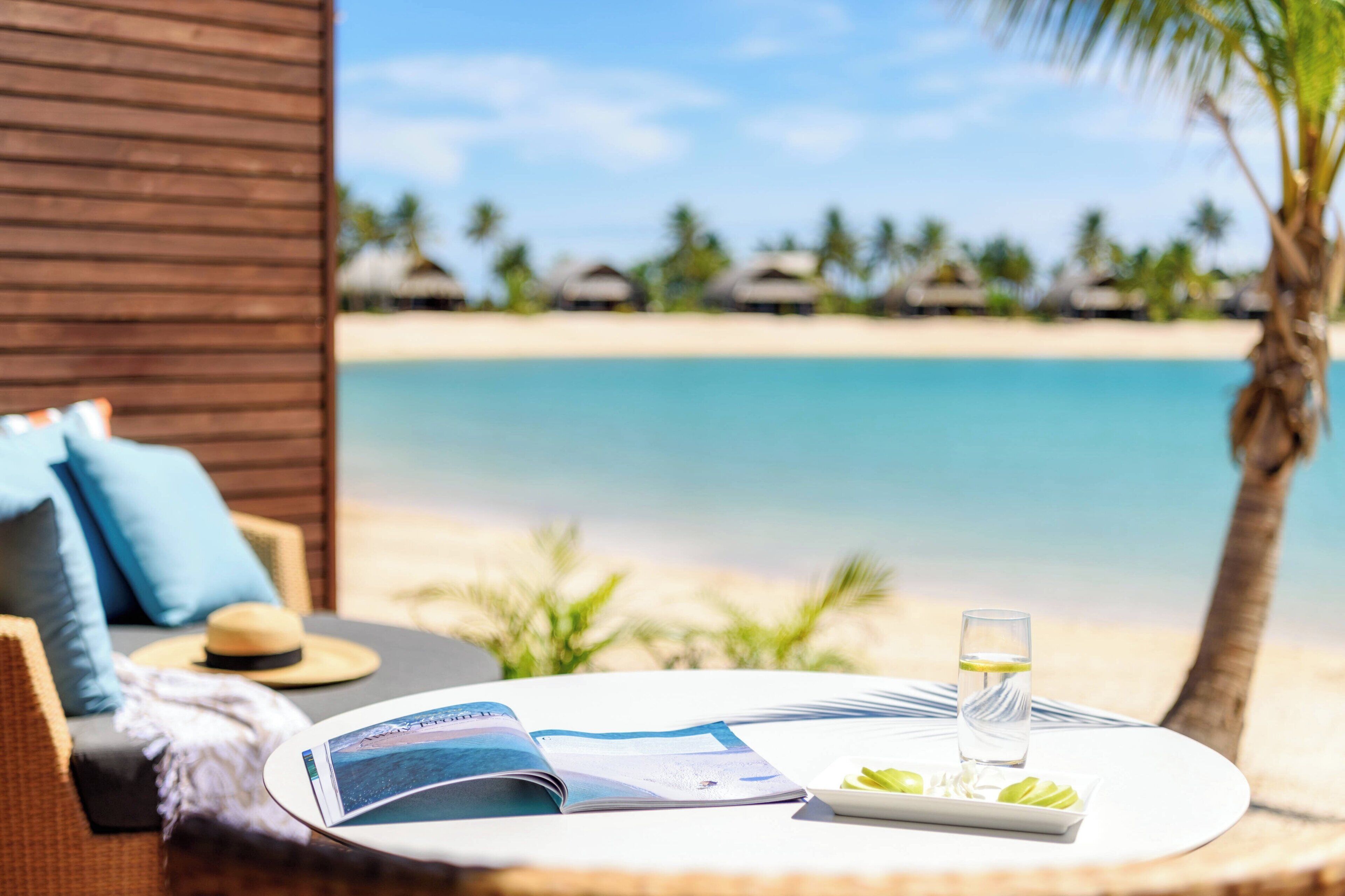Un área de descanso en la playa con vista al océano, una revista, un vaso de agua y un plato de fruta.